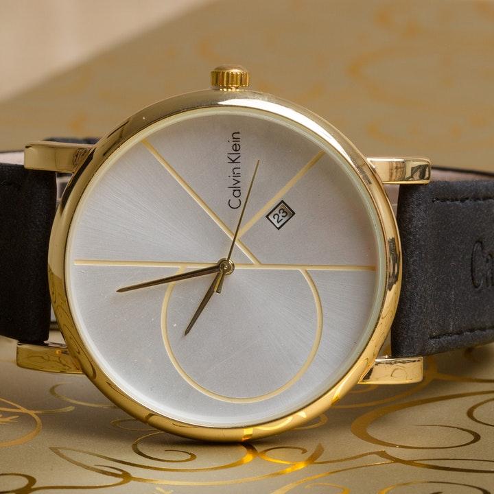 A close-up of a Calvin Klein wristwatch with a golden case finish, sleek gray dial, and navy blue leather band on a patterned background. The watch has a date indicator at the 3 o'clock position.