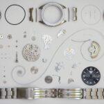 An array of disassembled wristwatch parts laid out neatly on a white background, including a watch face, various gears, springs, screws, and a metal band.