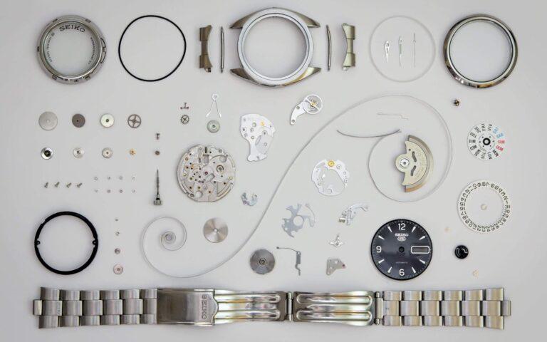 An array of disassembled wristwatch parts and tools laid out neatly on a white background, including a watch face, various gears, springs, screws, and a metal band.