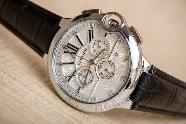 A close-up of an Cartier wristwatch with a cabochon crown, a silver case, white dial, multiple subdials, and a black leather strap, placed on a beige textured background.