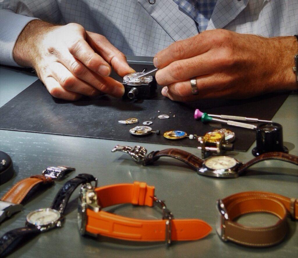 A close-up of a watchmaker's hands as he performs watch maintenance, surrounded by various watch parts and tools on a desk.