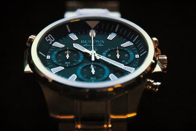 A close-up of a Bulova wristwatch with a gleaming silver case and dark teal face, featuring white markings and hands, set against a black background.