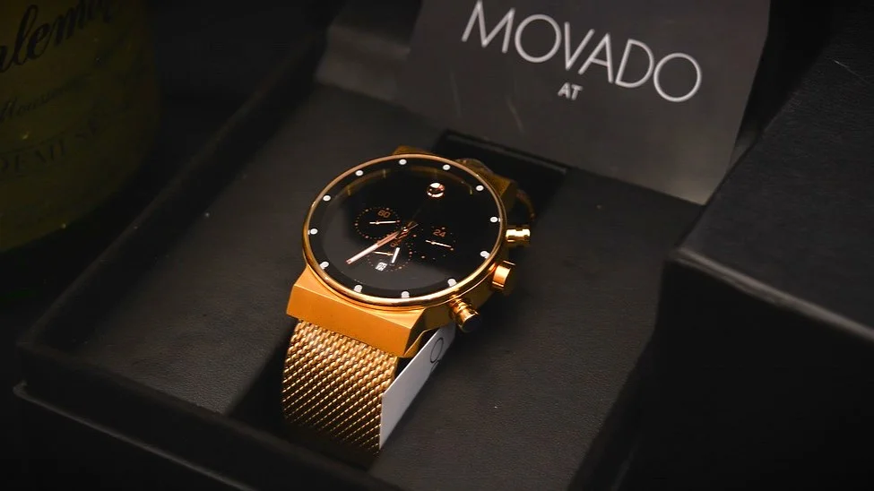 A gold-tone Movado watch with a Milanese bracelet is placed inside a box.