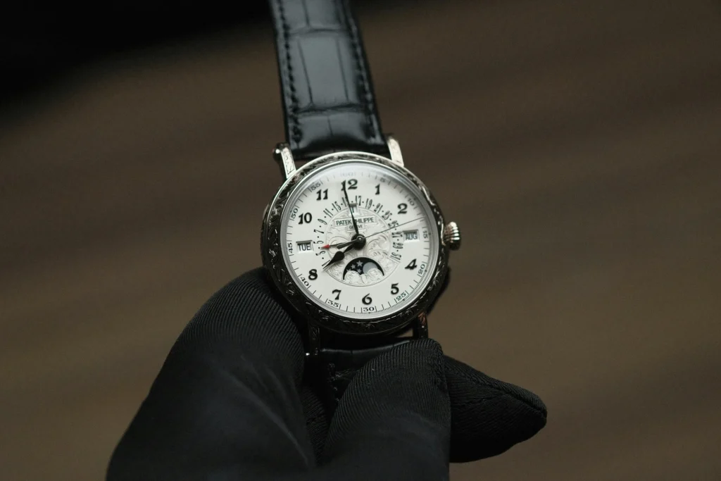 A person with black gloves, holding a Patek Philippe automatic watch.