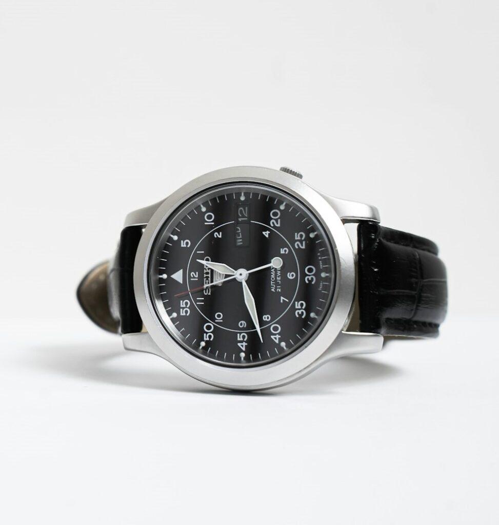 An analog Seiko watch with a stainless steel case, black leather strap, and black dial featuring large contrasting Arabic numerals and white hands.