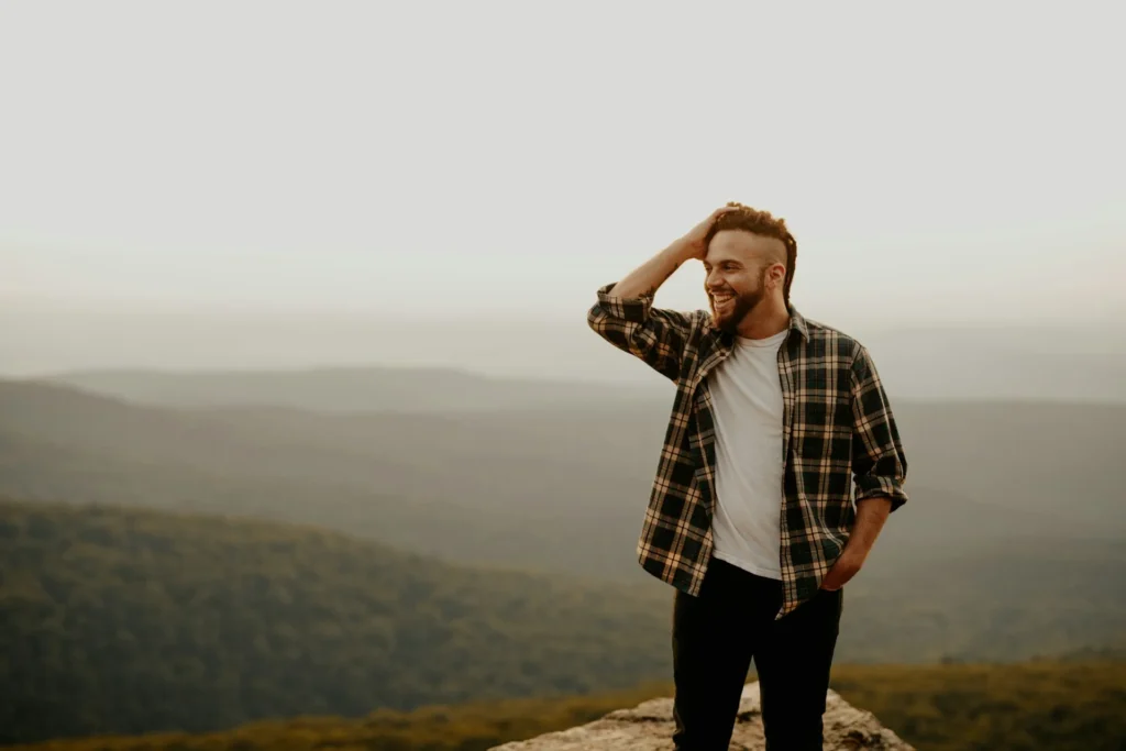 A man in a dress shirt and jeans stands atop a mountain, hands on his head, enjoying the breathtaking view.