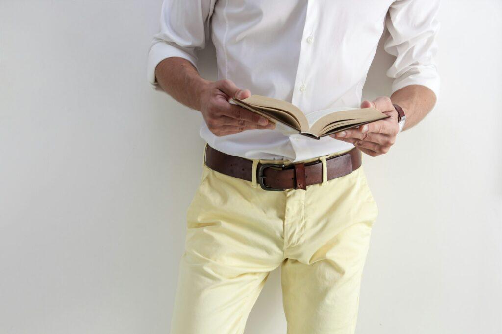  A man dressed in yellow pants and a white shirt, holding a book, featuring a dark brown belt as part of his attire.