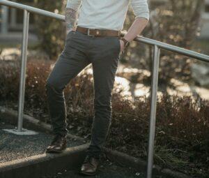 A tattooed man in a white shirt rests against a railing, featuring a coordinated belt and shoes for a stylish look.