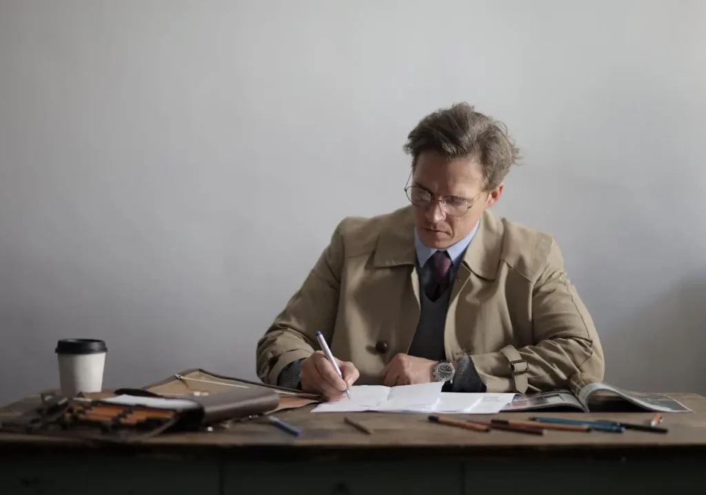 A man wearing a trench coat is seated at a desk, diligently writing with a pen on a sheet of paper.