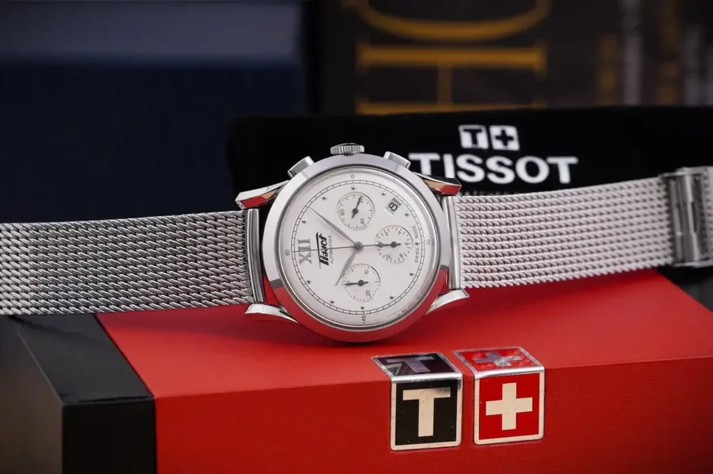 A Tissot Heritage watch with cream dial and mesh bracelet resting on it's box horizontaly