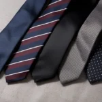 Five essential men's ties featuring royal blue grenadine, burgundy regimental striped, classic black silk, gray herringbone, and navy polka dot designs, arranged diagonally on a light gray linen background.